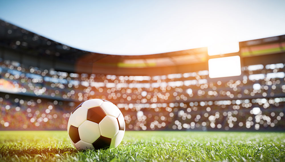Soccer ball on pitch with stadium in background.